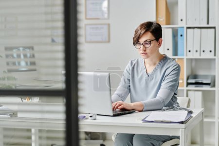 Foto de Young serious female physician or nurse consulting online patients in clinical office while sitting by workplace in front of laptop - Imagen libre de derechos