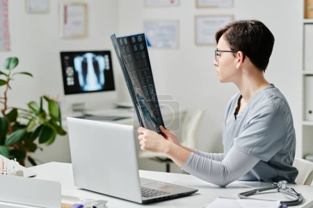 Téléchargez les photos : Young serious female clinician in blue medical scrubs learning x-ray image of patient during online consultation by workplace in office - en image libre de droit