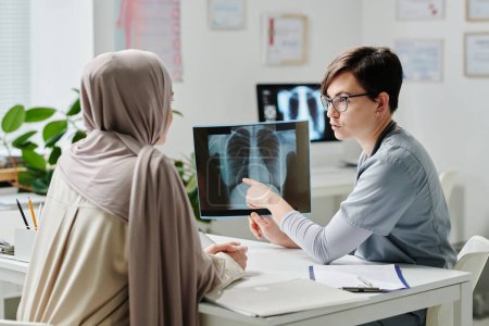 Téléchargez les photos : Radiologiste confiante montrant le résultat de l'examen médical à la patiente en hijab alors qu'elle était assise à son bureau devant elle - en image libre de droit