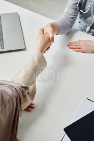 Foto de Young female clinician in blue uniform and Muslim patient shaking hands over desk with laptop and medical documents in hospital - Imagen libre de derechos