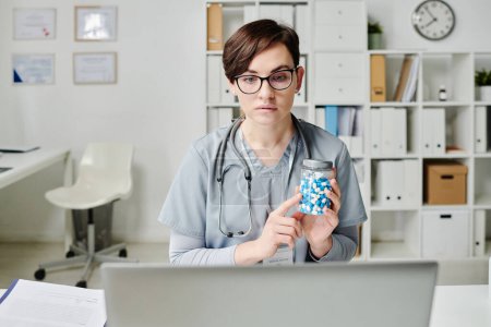 Téléchargez les photos : Young serious doctor showing jar of pills to patient during online consultation while sitting in front of laptop in medical office - en image libre de droit