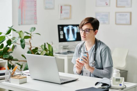 Téléchargez les photos : Contemporary young female clinician with jar of pills consulting online patient in office while sitting in front of laptop by desk - en image libre de droit