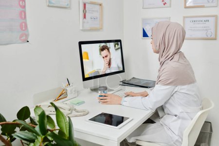 Téléchargez les photos : Young Muslim female doctor in hijab consulting online male patient with covid coughing or sneezing in front of computer screen - en image libre de droit