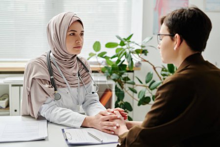Photo for Young clinician in hijab looking at sick and upset female patient sitting by desk in front of her in medical office during consultation - Royalty Free Image