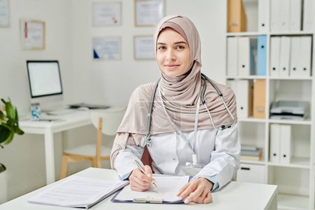 Foto de Joven doctora exitosa en hijab y whitecoat mirando la cámara mientras está sentada en el lugar de trabajo y rellenando la forma médica del paciente - Imagen libre de derechos
