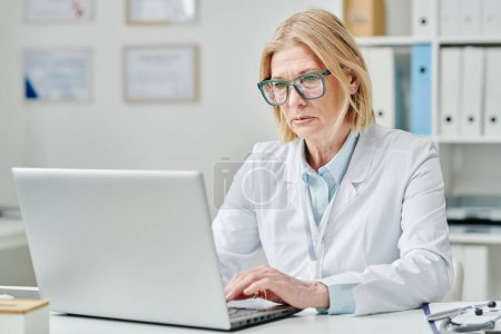 Téléchargez les photos : Serious mature blond female doctor in whitecoat and eyeglasses typing on laptop keyboard while sitting by desk in medical office - en image libre de droit