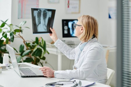 Téléchargez les photos : Radiologiste blonde en blanc regardant l'image radiographique des poumons des patients assis devant un ordinateur portable pendant la consultation en ligne - en image libre de droit