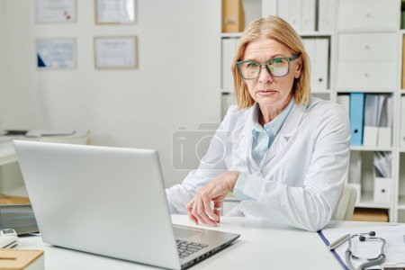Photo for Confident mature experienced doctor in whitecoat sitting by workplace in front of laptop and looking at camera during online consultation - Royalty Free Image
