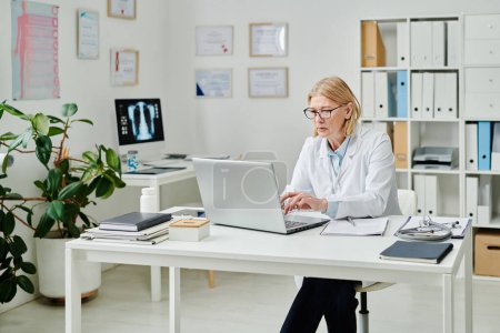 Foto de Serious mature female clinician in whitecoat answering questions of online patients while typing on laptop keyboard by desk - Imagen libre de derechos