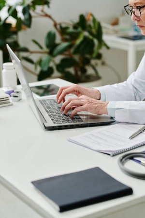 Téléchargez les photos : Close-up of mature experienced clinician concentrating on consulting online patients and answering their questions in medical office - en image libre de droit