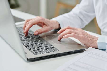 Photo for Hands of mature female clinician pressing buttons of laptop keyboard while sitting by desk and consulting patients online - Royalty Free Image