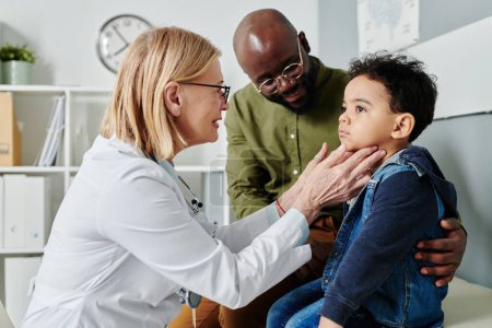 Photo for Experienced emale endocrinologist touching neck of little child while checking his thyroid during visit to clinics with his father - Royalty Free Image