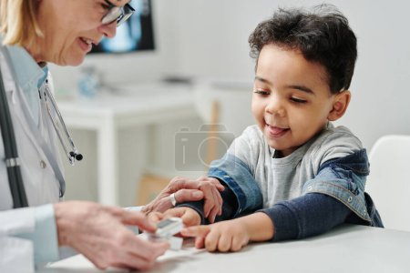 Téléchargez les photos : Contemporary pediatrician putting pulse oximeter on fingertip of cute little patient to check level of oxygen saturation in his blood - en image libre de droit