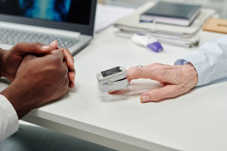 Photo for Hand of mature female patient with pulse oximeter on fingertip sitting in front of doctor during check-up of oxygen level in blood - Royalty Free Image