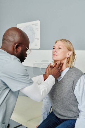 Téléchargez les photos : African American male endocrinologist checking thyroid of mature female patient in casualwear sitting by wall in front of him in hospital - en image libre de droit