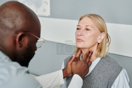 Téléchargez les photos : Femme blonde mature ayant un examen de ses glandes thyroïdes par un endocrinologue afro-américain dans un cabinet médical - en image libre de droit