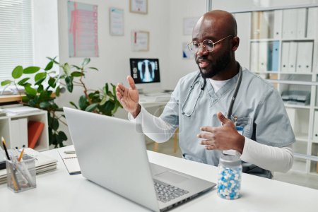 Téléchargez les photos : Young confident African American online doctor in uniform sitting by desk in front of laptop in clinics and giving medical advice to patient - en image libre de droit