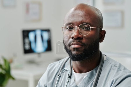 Téléchargez les photos : Young serious African American male general practitioner in blue uniform and eyeglasses sitting by workplace in front of camera in hospital - en image libre de droit