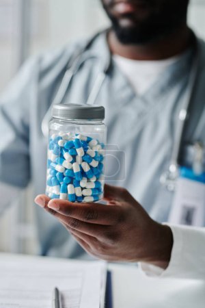 Photo for Big transparent plastic jar with pills held by contemporary male clinician in blue uniform sitting in front of camera in medical office - Royalty Free Image