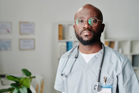 Photo for Young contemporary African American male clinician in eyeglasses and blue uniform standing in medical office and looking at camera - Royalty Free Image