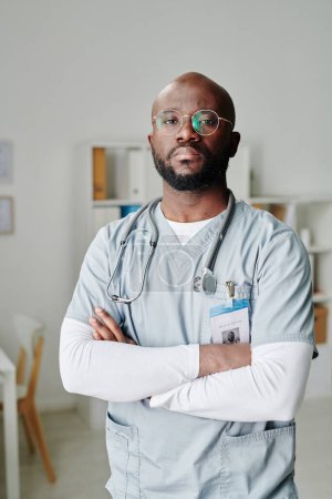 Foto de Grave joven afroamericano clínico con fonendoscopio en el cuello cruzando los brazos por el pecho mientras está de pie en el consultorio médico - Imagen libre de derechos