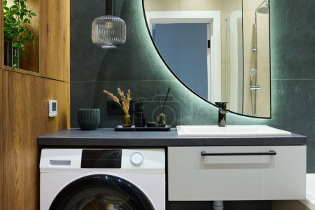 Téléchargez les photos : Bougie parfumée, désodorisant roseau et bouteille de remplissage en verre noir sur la machine à laver à côté de l'évier avec lampe et miroir demi-cercle ci-dessus - en image libre de droit