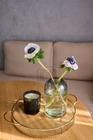 Photo for Two wildflowers with long stems standing in jar with water next to scented candle in black glass in round handmade wicker tray on table - Royalty Free Image