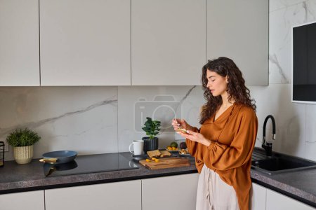 Téléchargez les photos : Jeune femme en bonne santé en chemisier et pantalon étaler le fromage sur une tranche de pain tout en faisant un sandwich pour le petit déjeuner dans la cuisine - en image libre de droit