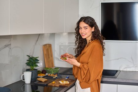 Foto de Feliz joven mujer contemporánea con sándwich vegetariano mirando a la cámara mientras está de pie en la cocina y desayunando - Imagen libre de derechos