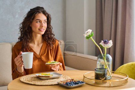 Photo for Happy young female vegetarian in casual blouse having tea with sandwich for breakfast while sitting by kitchen table in the morning - Royalty Free Image