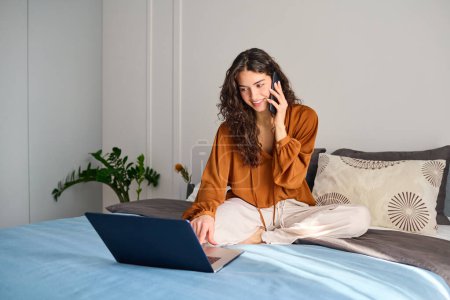 Foto de Young busy female in homewear looking at laptop screen and speaking on mobile phone while ordering something in online shop - Imagen libre de derechos
