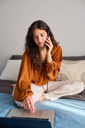 Photo for Pretty brunette girl in home wear scrolling through online goods while ordering one or some of them during conversation with operator - Royalty Free Image