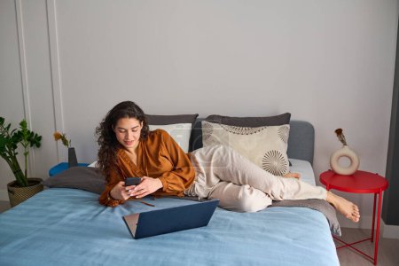 Photo for Young contemporary woman with smartphone relaxing on double bed in front of laptop while choosing goods during e-shopping - Royalty Free Image