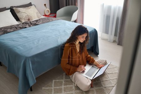 Photo for Busy young female with laptop sitting on the floor by double bed and networking or just spending free time on various websites - Royalty Free Image