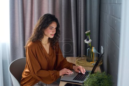 Photo for Young brunette woman in brown blouse sitting by desk in front of laptop and looking through online news or carrying out work - Royalty Free Image