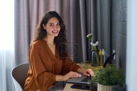 Téléchargez les photos : Jeune pigiste féminine réussie en chemisier brun travaillant sur ordinateur portable par lieu de travail dans le salon et regardant la caméra avec le sourire - en image libre de droit