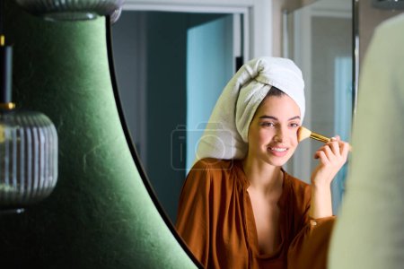 Téléchargez les photos : Reflection in mirror of happy young woman with white soft towel on head applying powder on her face with brush in the morning - en image libre de droit