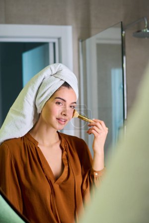 Téléchargez les photos : Contemporary young woman with towel on head applying makeup foundation on her face while standing in front of mirror - en image libre de droit