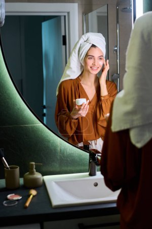Photo for Smiling young woman with white towel on head applying day cream on face in front of mirror while taking care of her beauty - Royalty Free Image