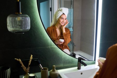 Téléchargez les photos : Fille gaie avec serviette blanche sur la tête choyer crème hydratante sur le visage après la douche du matin tout en regardant dans le miroir dans la salle de bain - en image libre de droit