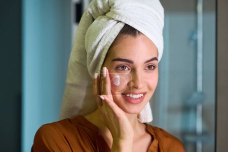 Photo for Reflection in mirror of healthy smiling girl with white soft towel on head pampering moisturizing cream on her face after bath - Royalty Free Image