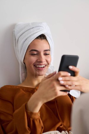 Téléchargez les photos : Young laughing female looking at smartphone screen while messaging with friends or communicating in video chat after bath - en image libre de droit