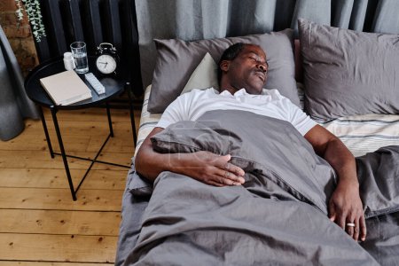 Téléchargez les photos : Homme âgé fatigué d'origine africaine dormant sous une couverture grise avec la tête sur un oreiller tout en se reposant sur un lit double confortable - en image libre de droit