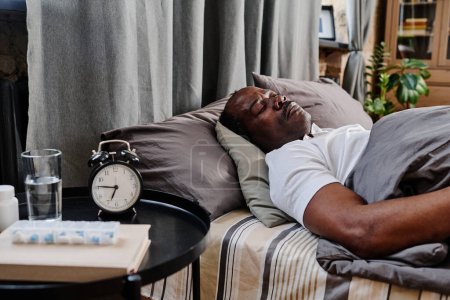 Téléchargez les photos : Aged African American man in white t-shirt sleeping under gray cover on comfortable double bed by small round table with alarm clock - en image libre de droit