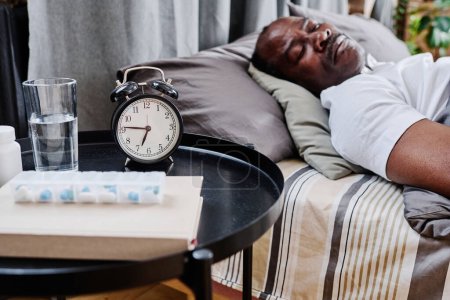Photo for Small round black table with alarm clock, medicaments, book and glass of water standing by double bed where senior man sleeping - Royalty Free Image