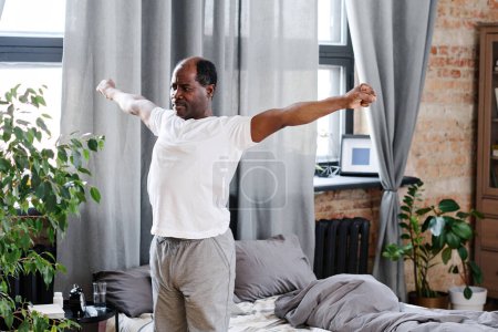 Téléchargez les photos : Homme noir âgé actif avec les bras tendus s'exerçant le matin tout en se tenant près de son lit avec des oreillers gris et une couverture - en image libre de droit