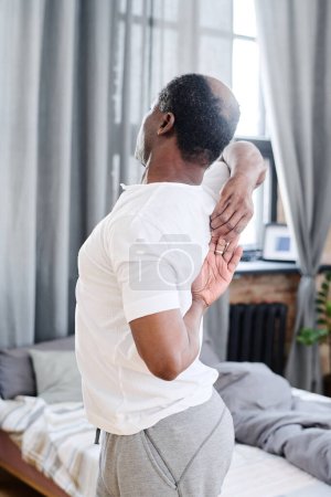 Téléchargez les photos : Afro-Américain âgé en pyjama faire de l'exercice d'étirement pour la moelle épinière tout en se tenant contre le lit devant la caméra - en image libre de droit