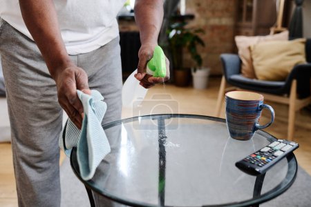 Foto de Hands of retired man with duster and detergent cleaning small glass round table with mug and remote control in living room - Imagen libre de derechos