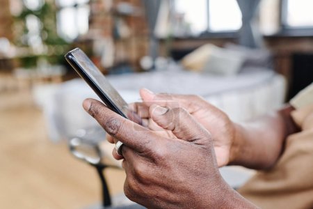 Téléchargez les photos : Les mains de l'homme noir retraité contemporain tenant téléphone portable et textos à temps libre dans sa chambre pendant la routine quotidienne - en image libre de droit