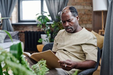 Téléchargez les photos : Homme retraité afro-américain contemporain assis dans un fauteuil et lisant un livre curieux à loisir tout en restant à la maison - en image libre de droit